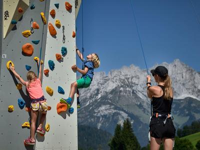 Dos niños escalan una pared de escalada al aire libre con vista a las montañas, entrenadora los observa.