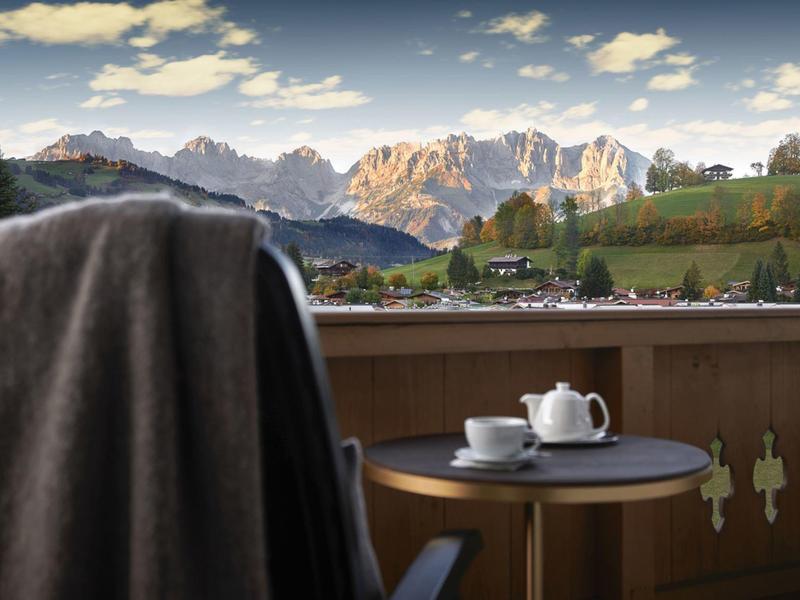 Balcón con mesa, tetera y taza, vista a colinas verdes y montañas iluminadas por el sol.