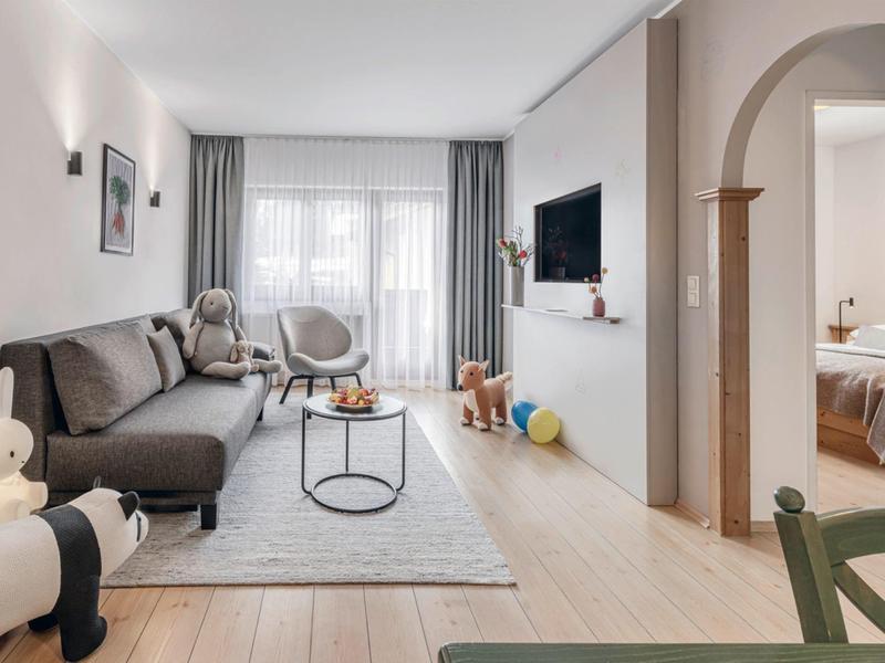 Sala de estar luminosa con sofá gris, alfombra, TV y acceso a un dormitorio.