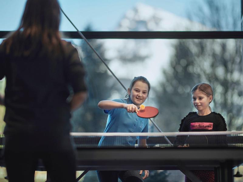 Tres personas juegan al tenis de mesa en una habitación con gran ventana y vista a la montaña.