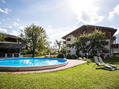 Okrągły basen na świeżym powietrzu na zielonym trawniku z leżakami przed dwupiętrowym budynkiem hotelu.