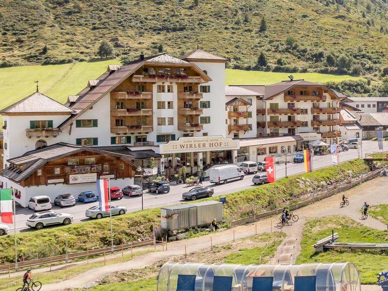 Groot hotelgebouw met houten gevel voor beboste heuvels en parkeerplaats.