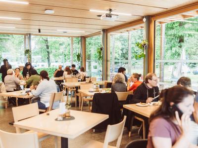 Interior of a café with guests at wooden tables and large windows overlooking greenery.