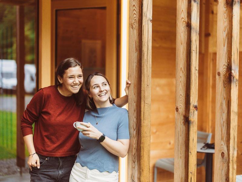 Two women lean against a wooden wall, smiling outside a rustic building.