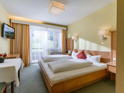 Bright hotel room with double bed, desk, TV, and balcony with curtains.
