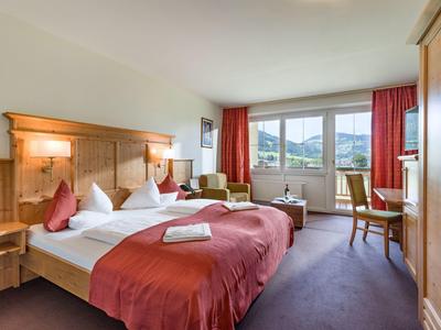 Bright hotel room with two double beds, red curtains, and mountain view through large windows.