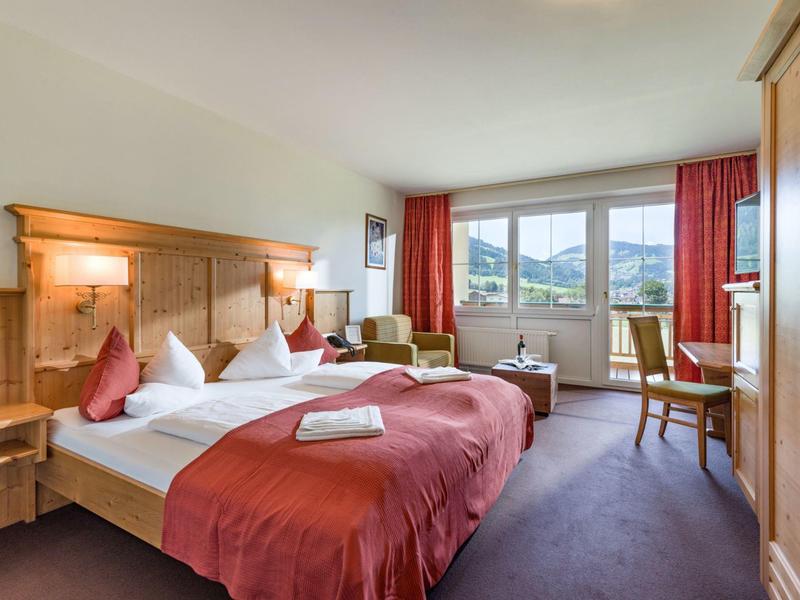 Bright hotel room with two double beds, red curtains, and mountain view through large windows.