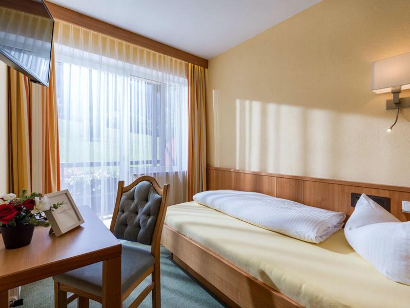 Bright hotel room with single bed, desk, chair, and window with sheer curtain.