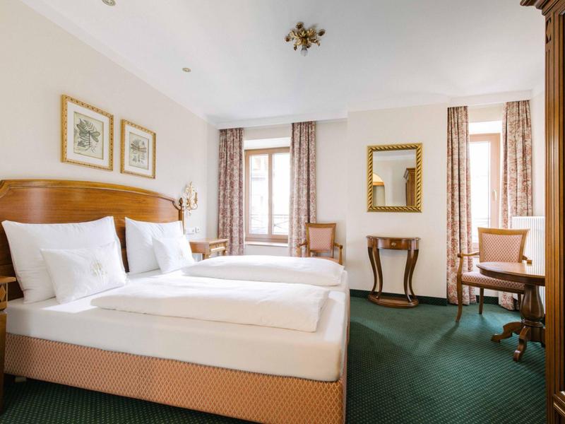 Camera d'hotel luminosa con letto matrimoniale, due poltrone e scrivania vicino a grandi finestre.