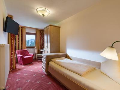 Kleine hotelkamer met eenpersoonsbed, fauteuils, kast en raam met gordijnen.