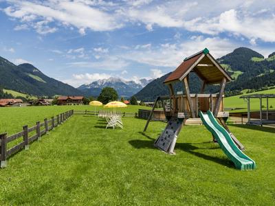Groot speelplein met glijbaan en klimrek op groen gras, omgeven door bergen.