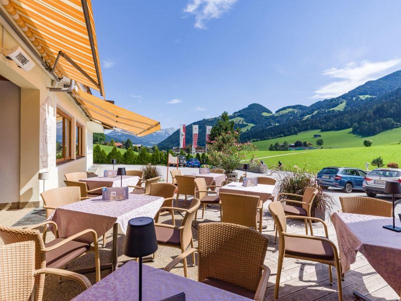 Zonnig terras met tafels en stoelen buiten een hotel met uitzicht op groene heuvels.