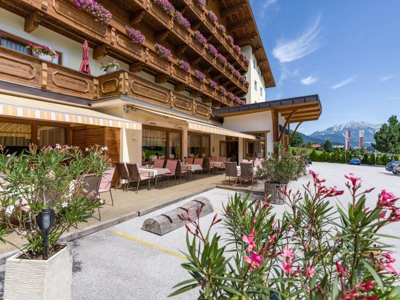 Buitenzijde van een hotel met terras, tafels en stoelen, omgeven door bloemen en bergen op de achtergrond.