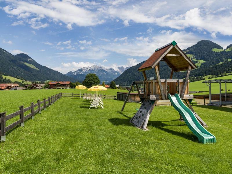 Groot speelplein met glijbaan en klimrek op groen gras, omgeven door bergen.