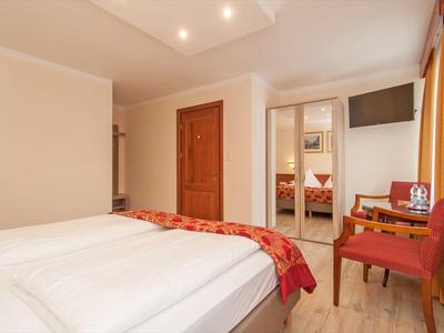 Jasny pokój hotelowy z łóżkiem podwójnym, czerwonym krzesłem i telewizorem na ścianie obok dużego okna z zasłonami.