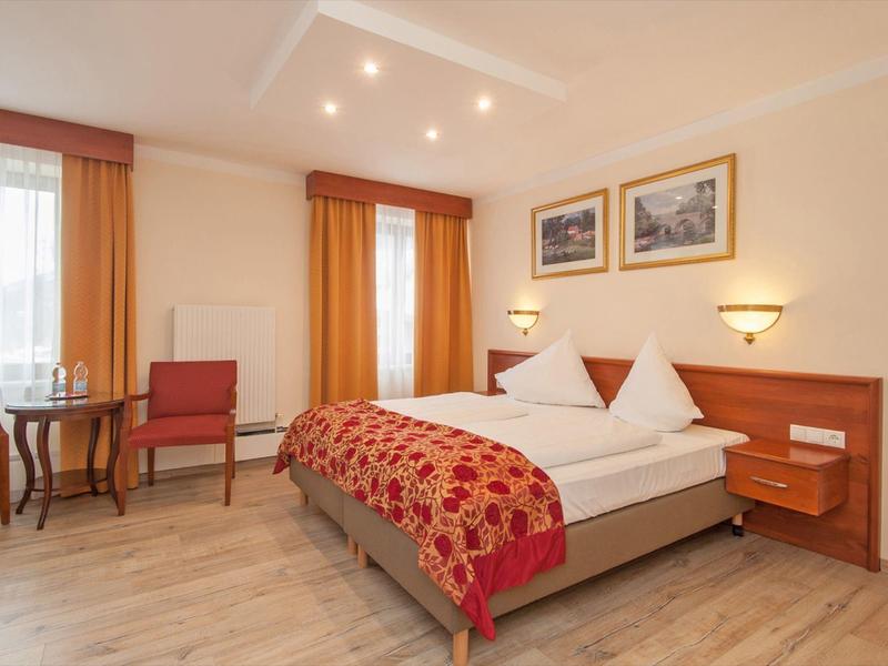 Bright hotel room with double bed, table, chairs, and two windows with orange curtains.