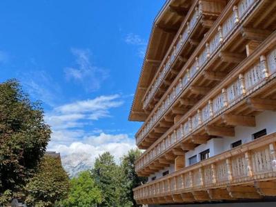 Mehrstöckiges Hotel mit Holzbalustraden neben einem gepflasterten Weg und Bäumen unter blauem Himmel.