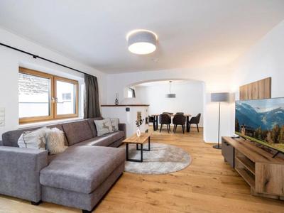 Helles Wohnzimmer mit grauem Sofa, Holzboden, TV und offenem Essbereich mit schwarz gepolsterten Stühlen.