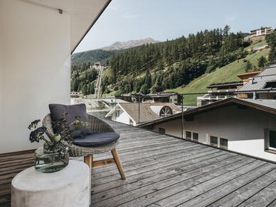 Balkon mit Holzboden, Stuhl und Blumen vor einer grünen Berglandschaft und Häusern.