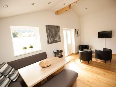 Heller, moderner Wohnbereich mit Holzboden, Sofa, Esstisch, zwei sesseln und Wandfernseher.