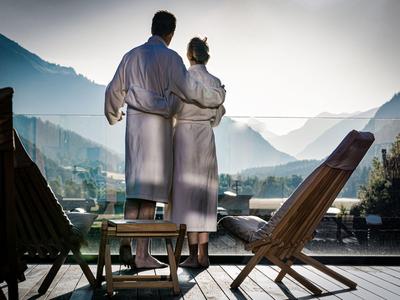 Couple en peignoirs admire le paysage montagneux depuis une terrasse en bois avec chaises longues.
