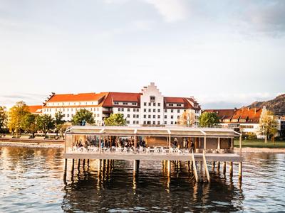 Historisches Hotelgebäude mit rotem Dach am Wasser, davor ein hölzerner Steg mit Sitzgästen.