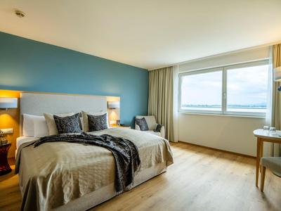 Helles Hotelzimmer mit großem Bett, blauer Wand und Fenstern mit Vorhängen.