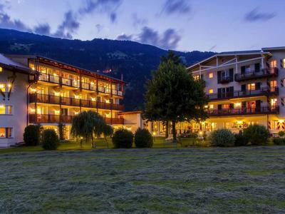 Beleuchtetes Hotelgebäude vor Bergkulisse bei Dämmerung mit grünem Vordergrund.