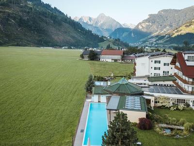 Hotel mit Außenpool zwischen grünen Bergen und einer weitläufigen Wiese unter klarem Himmel.
