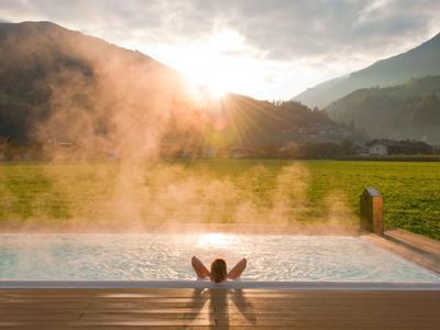 Person entspannt sich im Pool mit Blick auf sonnige Berge und grünes Feld im Morgenlicht.