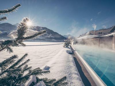 Winterlandschaft mit schneebedecktem See, Berge im Hintergrund und Dampfschwaden über einem Außenpool.