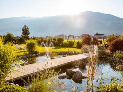 Sonniger Hotelgarten mit Teich, Holzbrücke, Springbrunnen und Bergblick im Hintergrund.