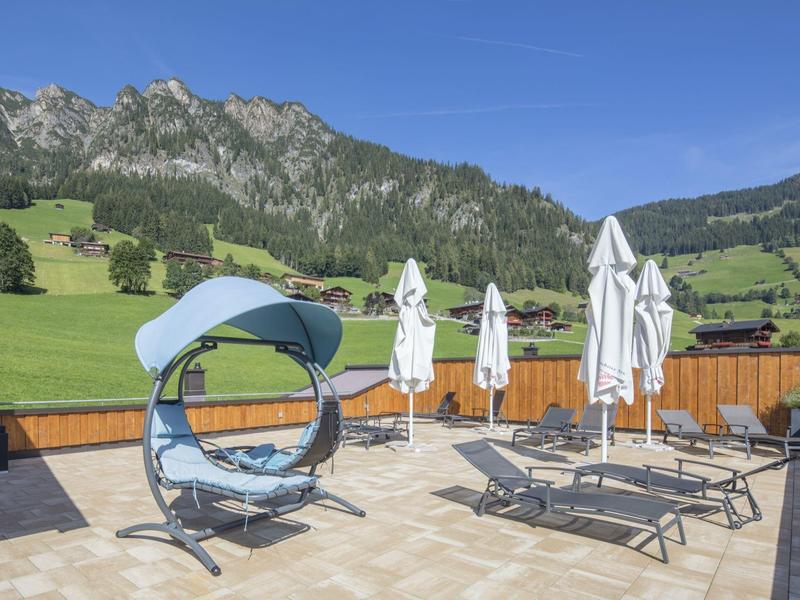 Terasa s moderními lehátky a závěsným křeslem před slunečnou horskou scenérií.