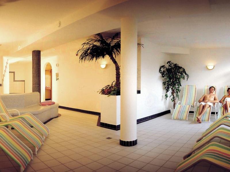 Innenraum eines Wellnessbereichs mit Liegestühlen, Säulen, Pflanzen und zwei Menschen am Whirlpool.