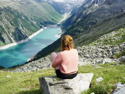 Frau in rosa Pullover sitzt auf Stein und blickt auf grünen Bergsee in Tal mit schneebedeckten Gipfeln.