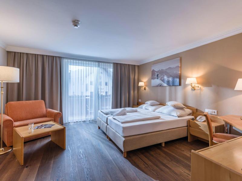 Helles Hotelzimmer mit Doppelbett, zwei Sesseln, Holzboden und großem Fenster mit Vorhängen.