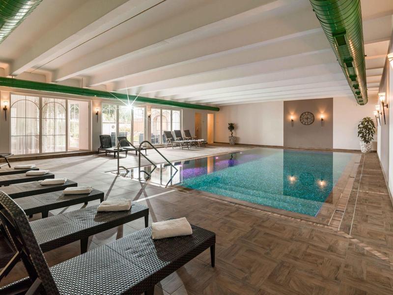 Innenpool mit Liegestühlen, großen Fenstern und Holzverkleidung, helle Decke mit grünen Akzenten.