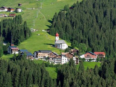 Kleines Dorf mit Kirche, Bauernhäusern und grünen Wiesen, umgeben von dichtem Fichtenwald.
