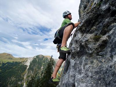 Mann klettert mit Helm und Rucksack an steiler Felswand in bergiger Landschaft bei bewölktem Himmel.