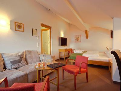 Gemütliches Zimmer mit Sofa, zwei roten Sesseln, Bett, TV und warmem Licht an weißer Wand.