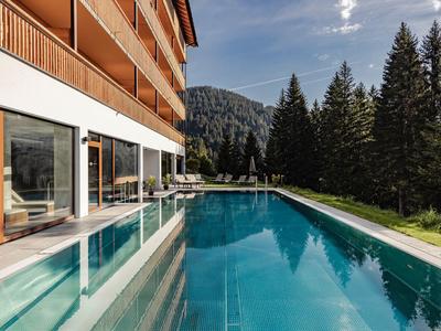 Modern outdoor pool beside a hotel with mountain and forest backdrop in sunny weather.