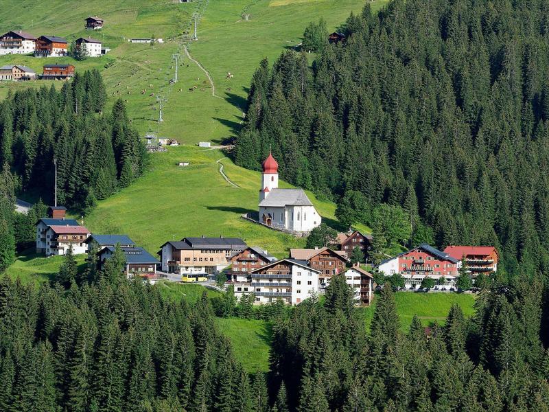 Kleines Dorf mit Kirche, Bauernhäusern und grünen Wiesen, umgeben von dichtem Fichtenwald.