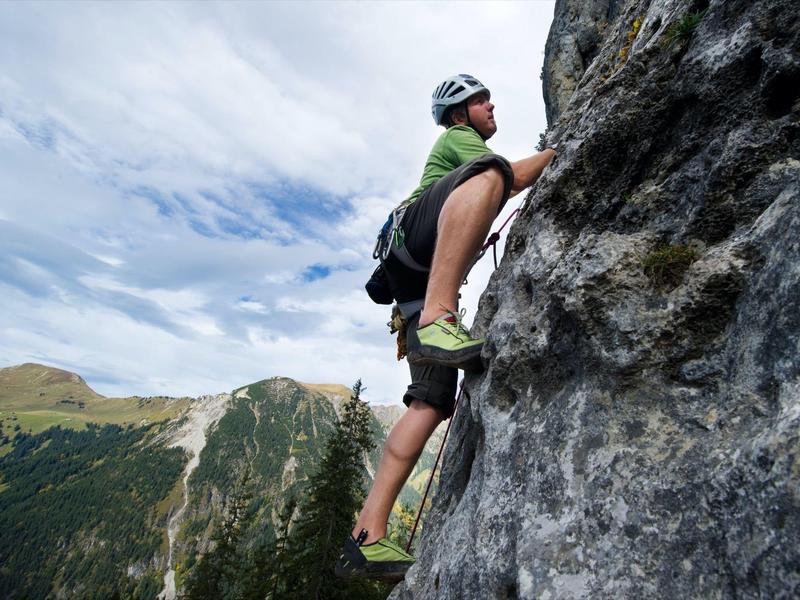 Mann klettert mit Helm und Rucksack an steiler Felswand in bergiger Landschaft bei bewölktem Himmel.
