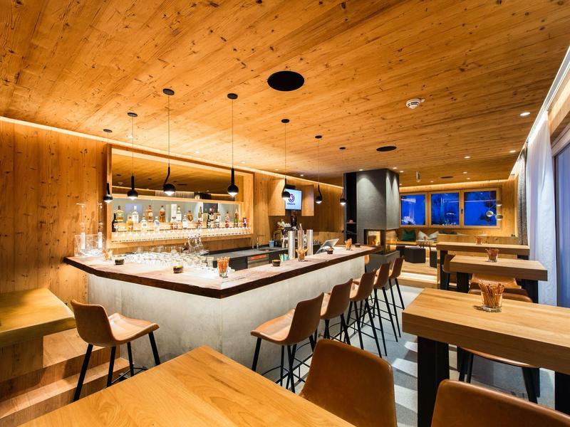 Moderner Barbereich mit Holzwänden, Bartresen, Hockern und Sitzgruppen, warme Beleuchtung, große Fenster.
