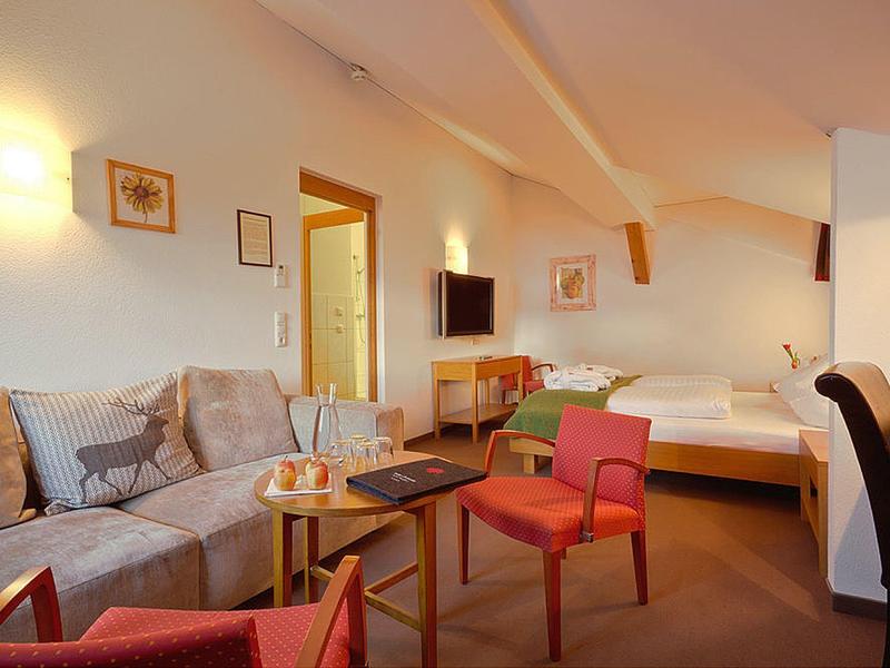 Gemütliches Zimmer mit Sofa, zwei roten Sesseln, Bett, TV und warmem Licht an weißer Wand.