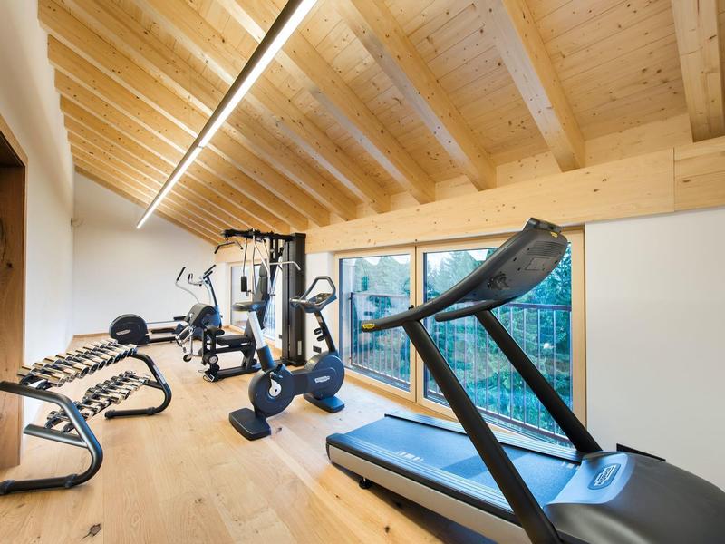 Moderner Fitnessraum mit Holzbalkendecke, Laufband, Kraftstation und Hanteln, großer Fensterblick.