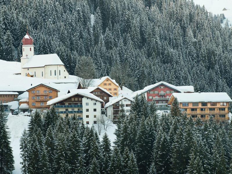 Winterliches Dorf mit verschneiten Dächern vor dichtem, bewaldetem Berghang.
