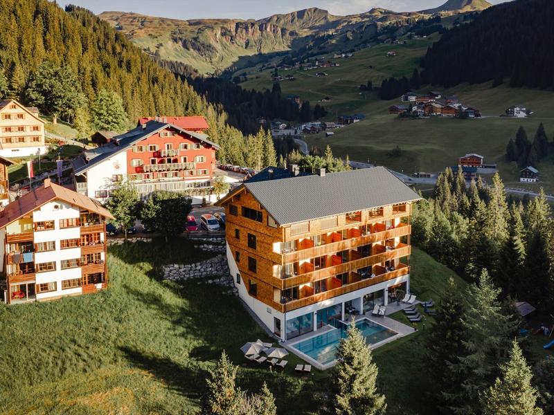 Berge mit Tannenwald umgeben mehrere mehrstöckige Holzgebäude und einen Pool in sonniger Landschaft.