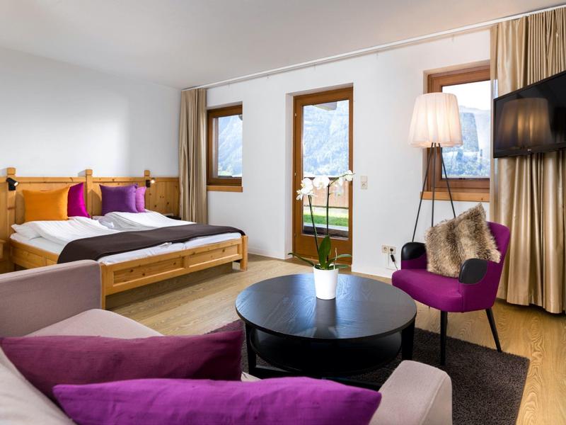 Gemütliches Zimmer mit Holzmöbeln, Doppelbett, lila Sofa, Stuhl, Tisch und Balkon mit Bergblick.