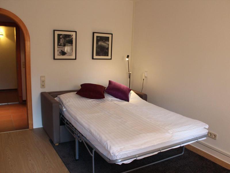 Schlafzimmer mit großem Bett, zwei dunklen Kissen, zwei Wandbildern und Stehlampe in der Ecke.